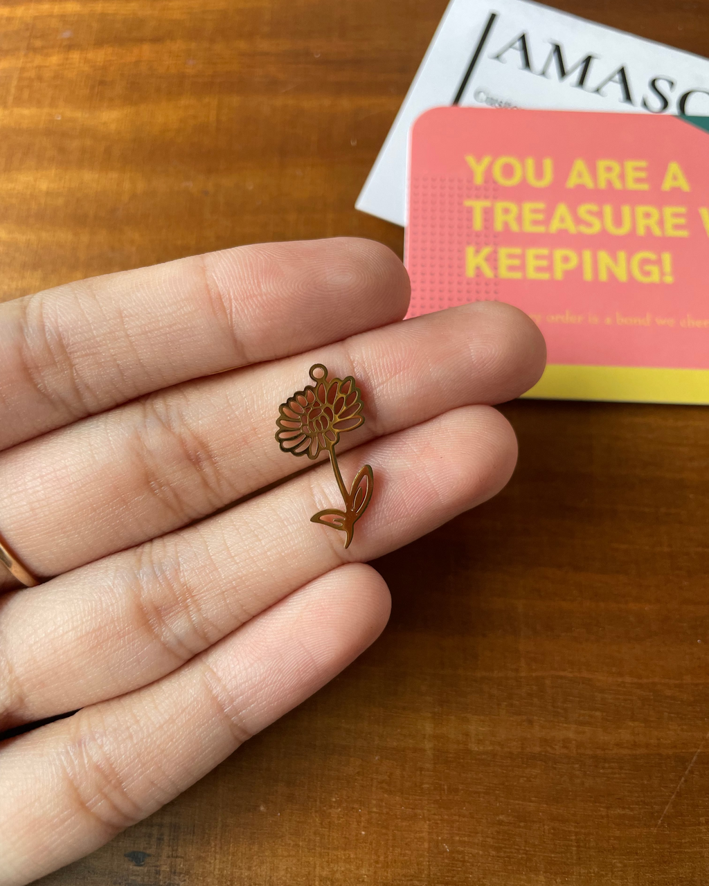 Hand holding a small gold flower-shaped object on a wooden surface with a card in the background.