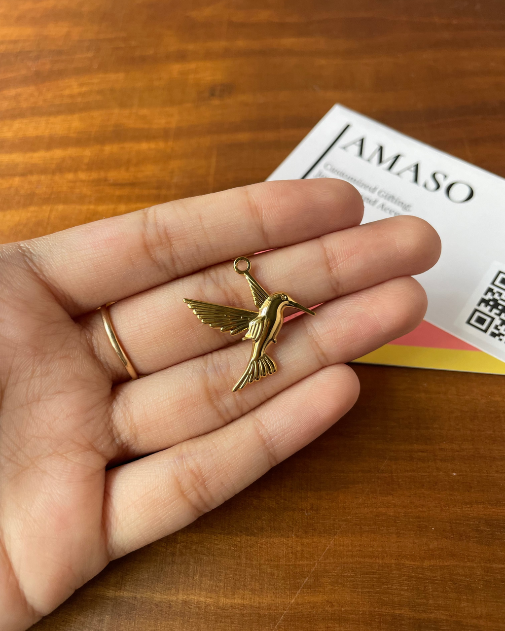 Gold hummingbird pendant held in a hand on a wooden surface with 'AMASO' branding.