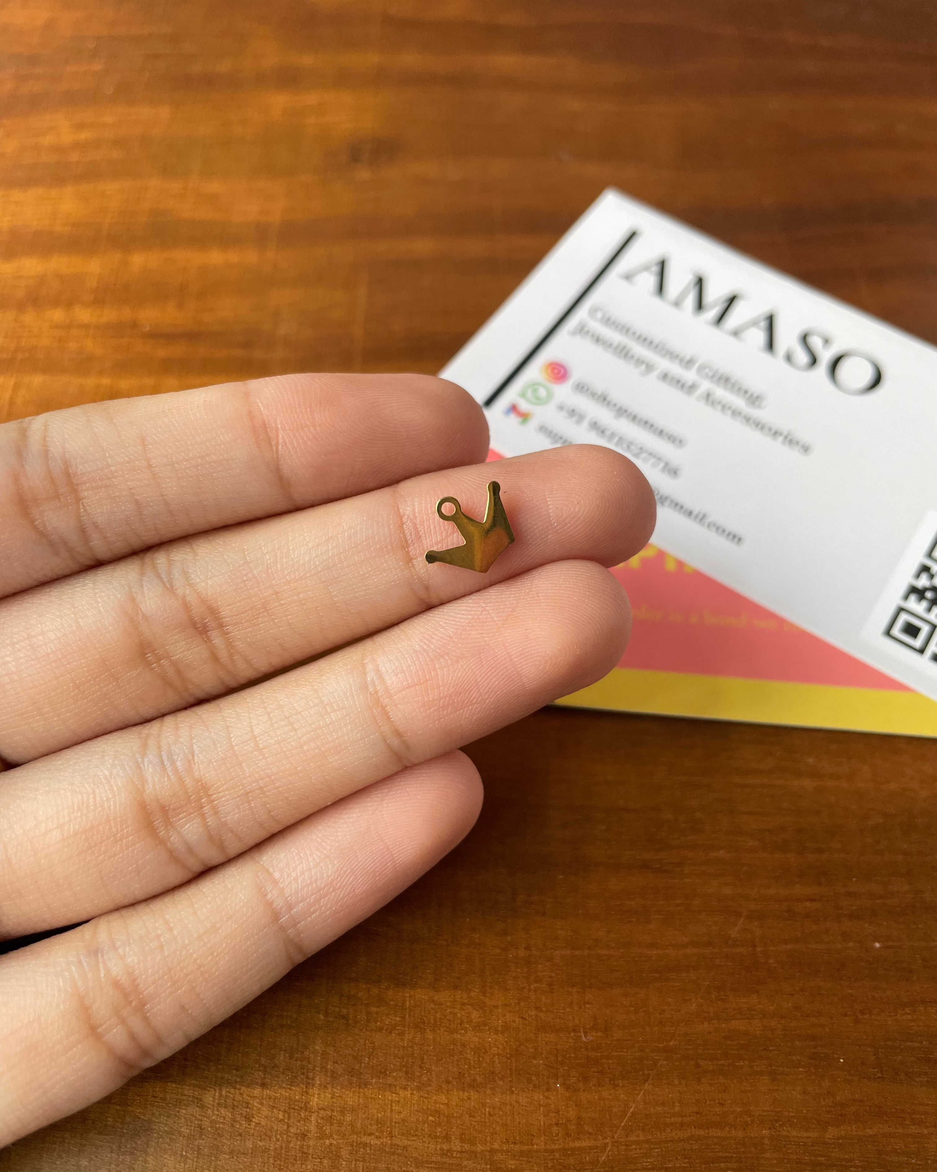Gold crown earring held between fingers on a wooden surface with AMASO card in the background