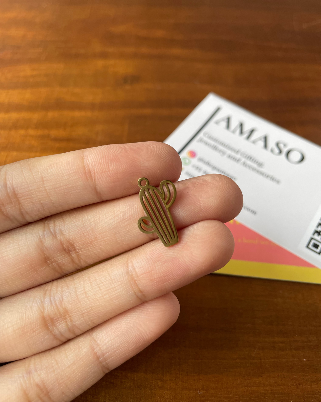 Small cactus-shaped object held between fingers on a wooden surface with AMASO branding in the background.