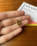 Gold teardrop pendant held in a hand with a wooden surface and AMASO card in the background