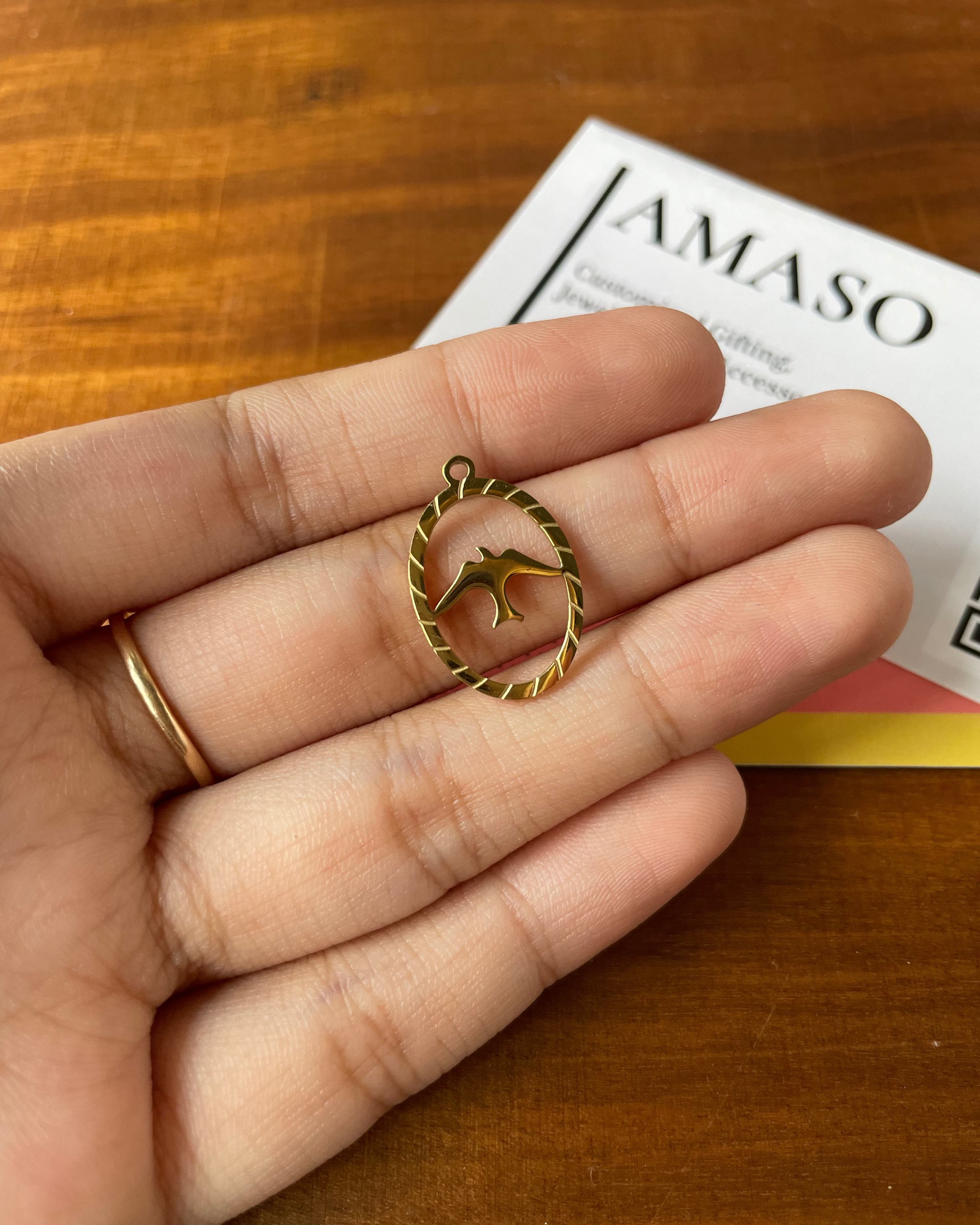 Gold pendant held in a hand with 'AMASO' card in the background