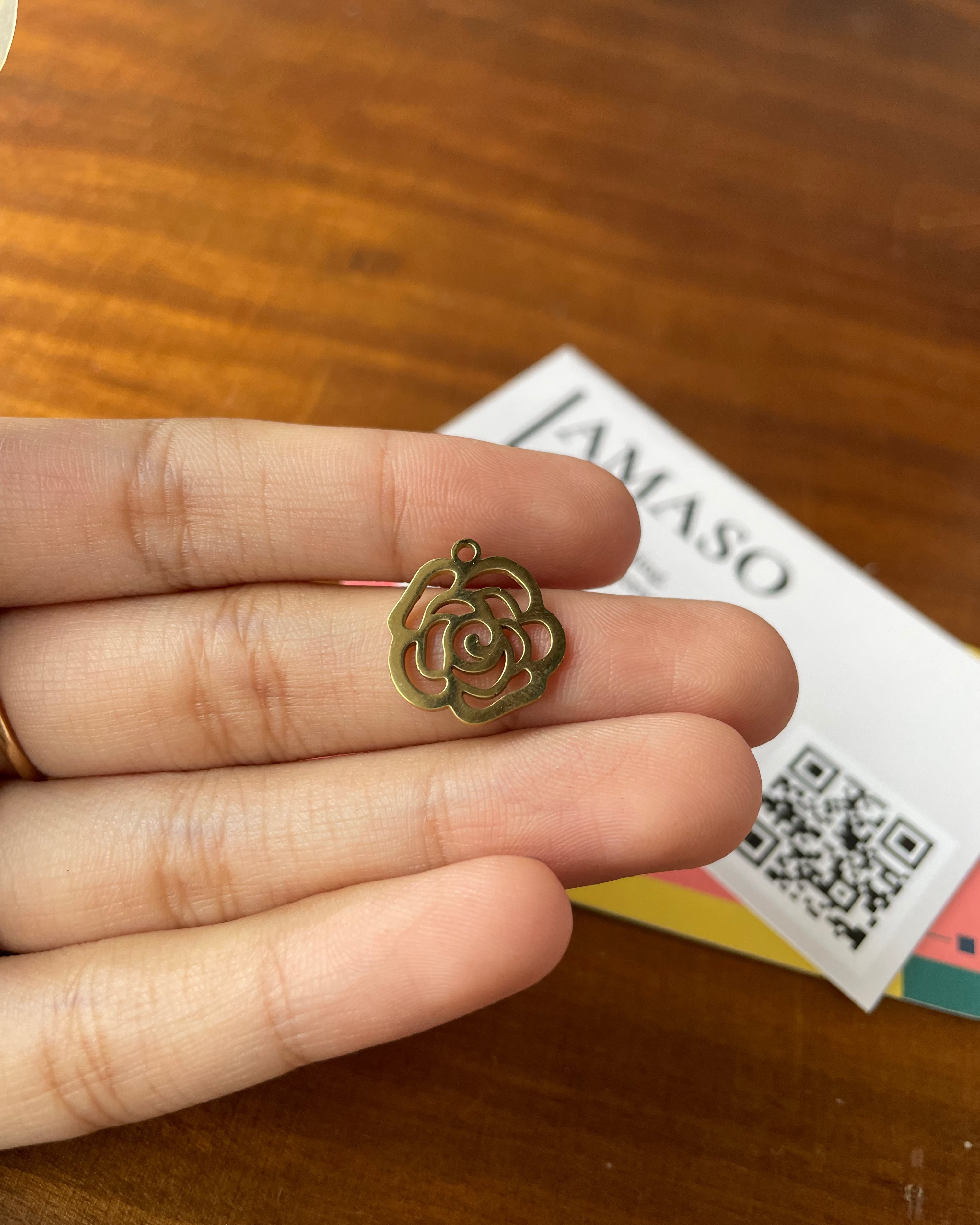 Gold rose-shaped pendant held between fingers on a wooden surface with a colorful card in the background.