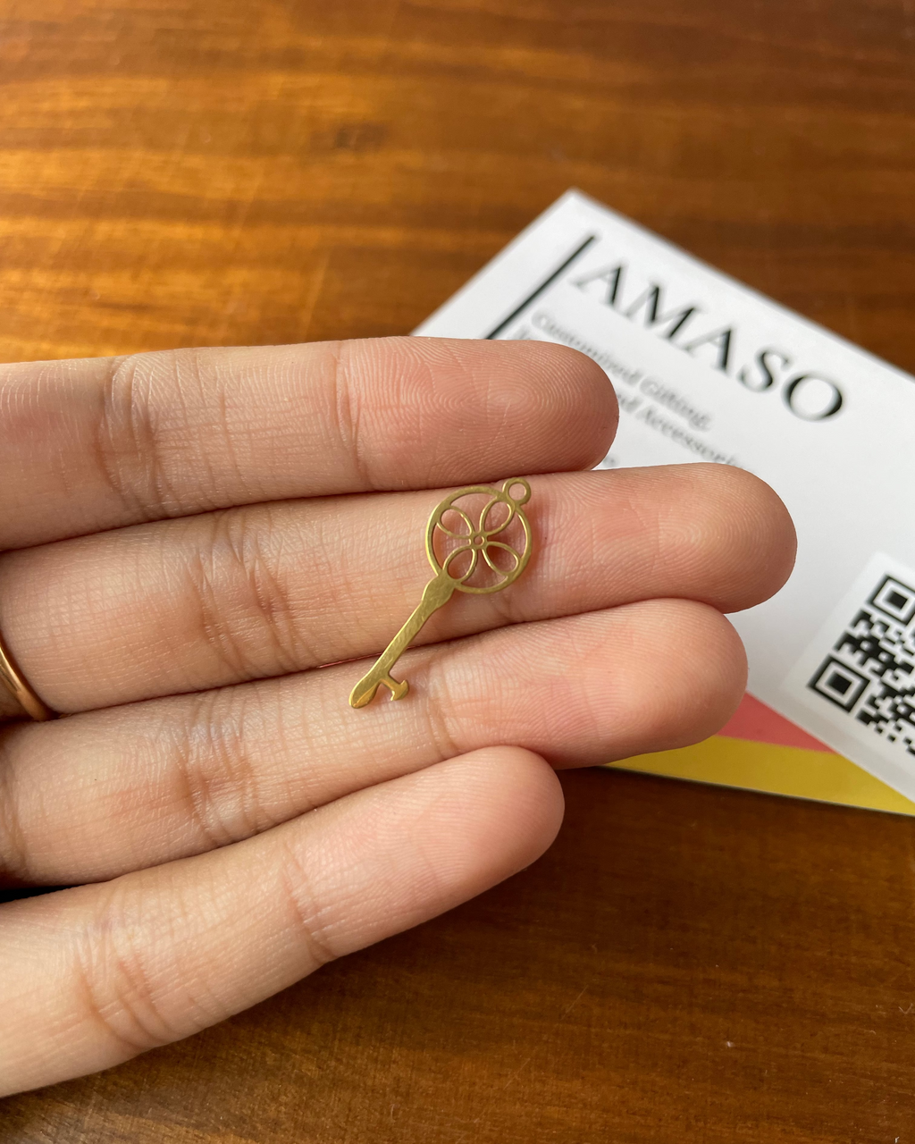 Gold key-shaped earring held between fingers on a wooden surface with 'AMASO' branding in the background.