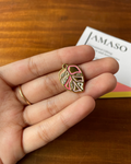 Hand holding a gold and pink leaf-shaped charm with AMASO packaging in the background.