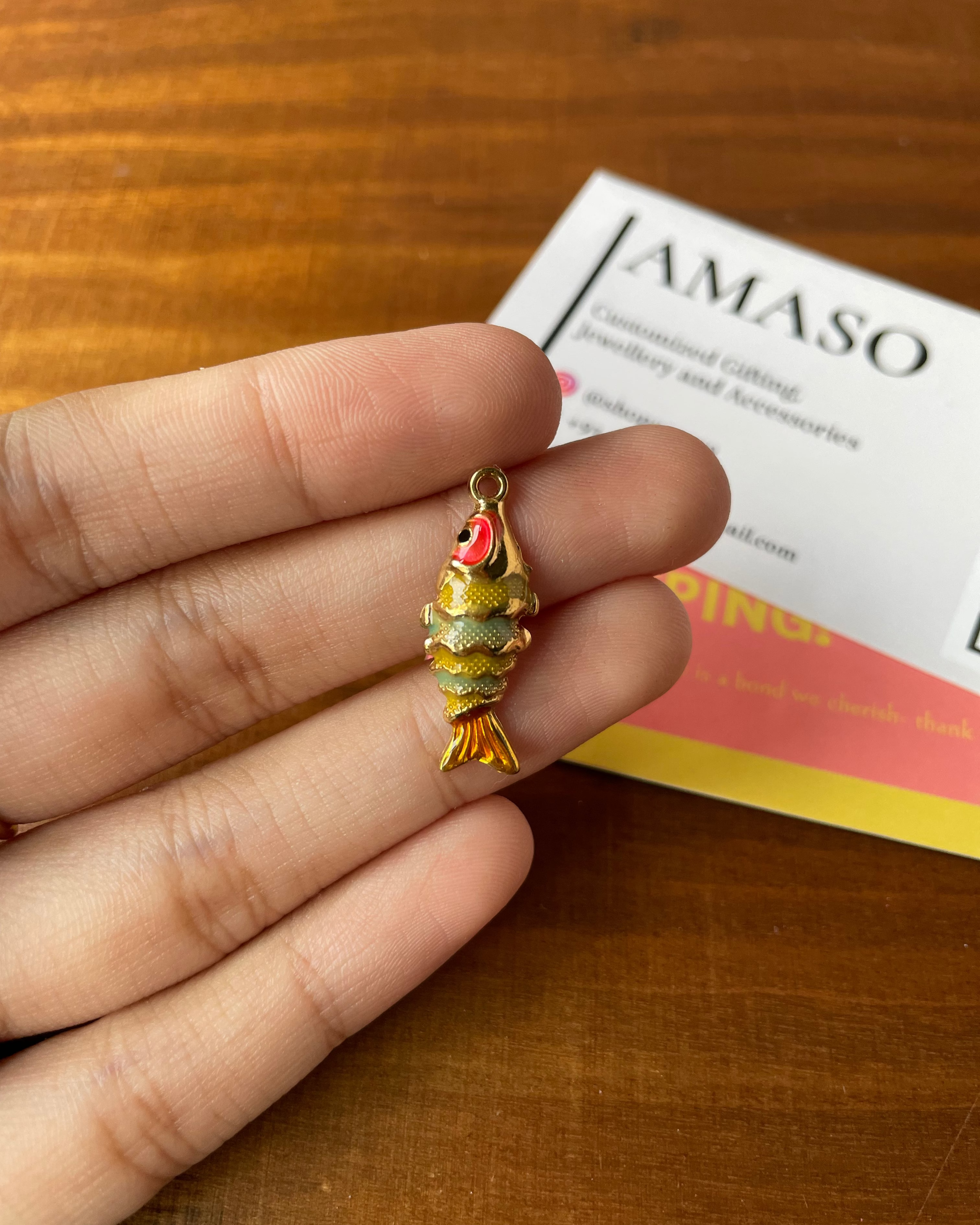Gold fish-shaped pendant held between fingers on a wooden surface with a card in the background.