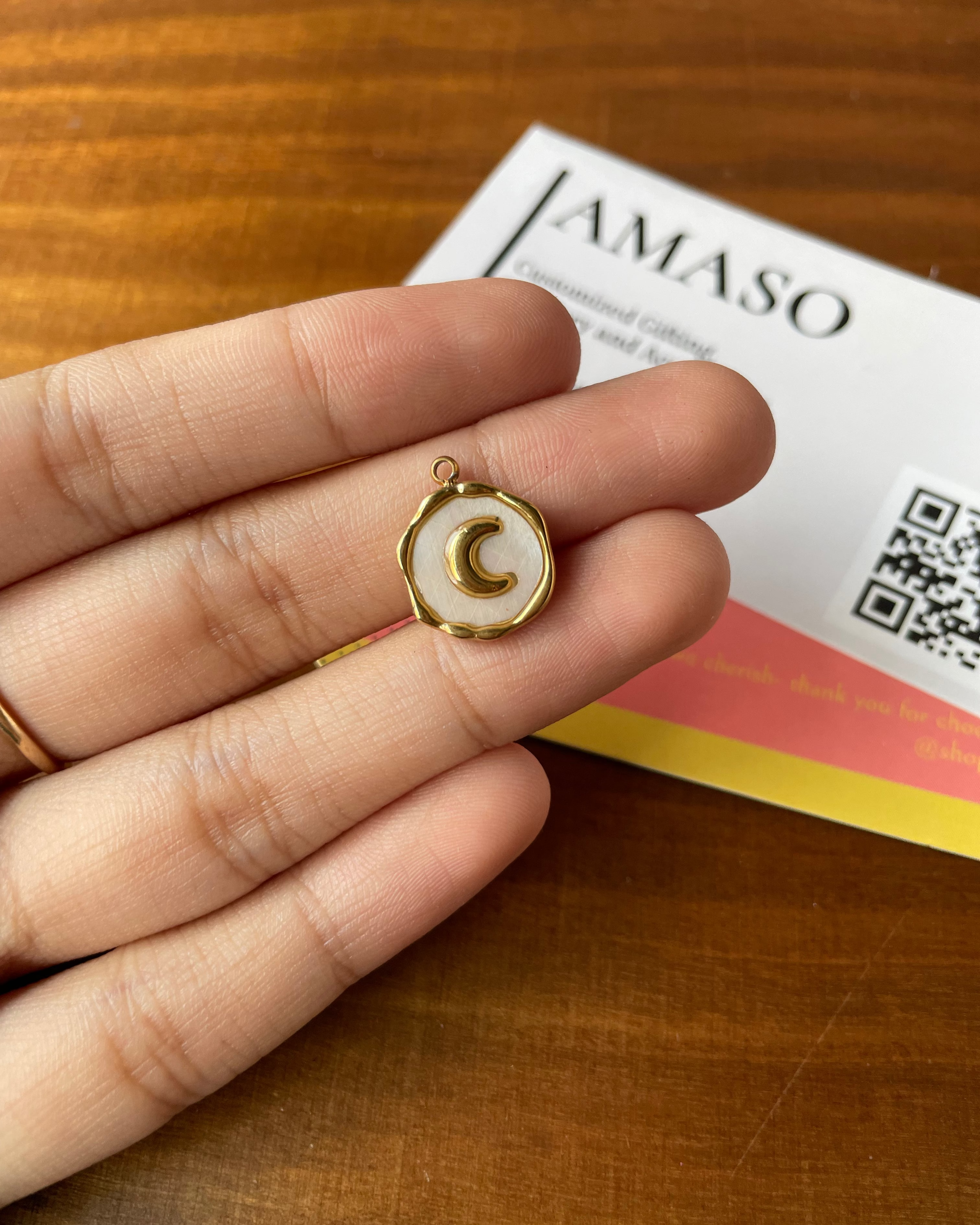 Gold and white pendant held between fingers with a 'JAMASO' product in the background.