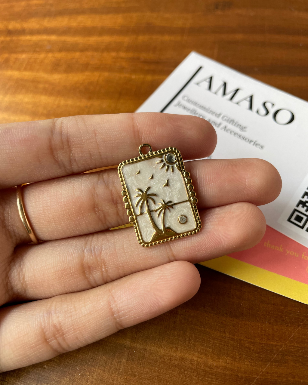 Hand holding a decorative pendant with palm tree design on a wooden surface with 'AMASO' card.