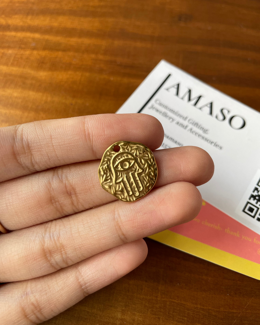 Gold pendant held in a hand with a 'AMASO' card in the background