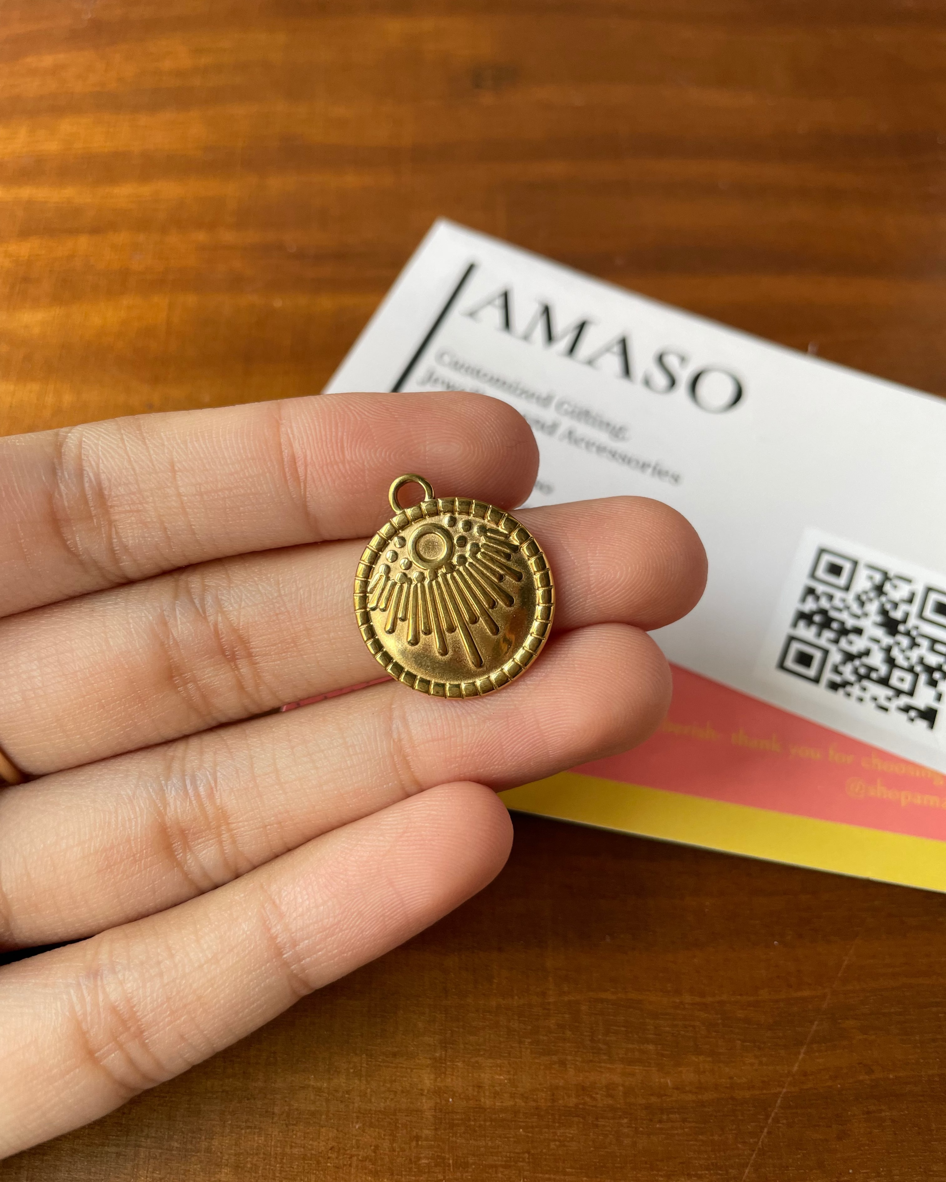 Gold pendant held in a hand with AMASO branded card in the background