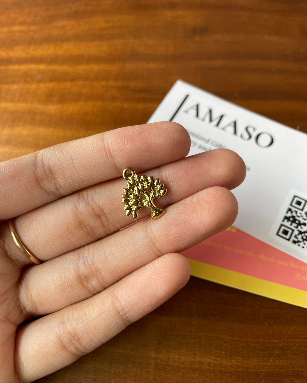 Gold tree-shaped pendant held between fingers with a 'JAMASO' card in the background.