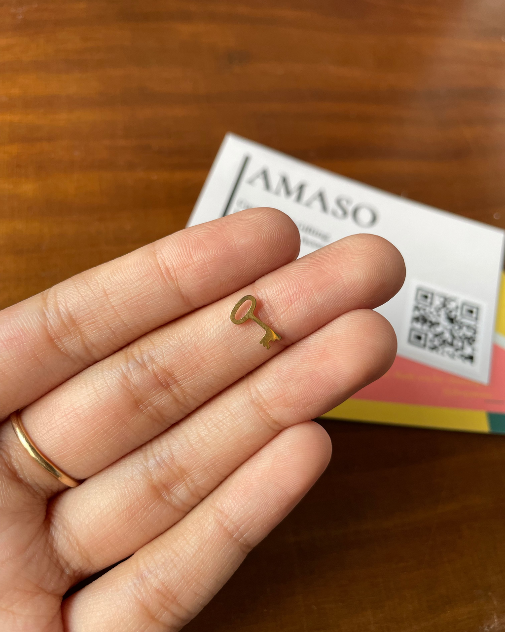 Gold key-shaped earring held between fingers on a wooden surface with a colorful box in the background.