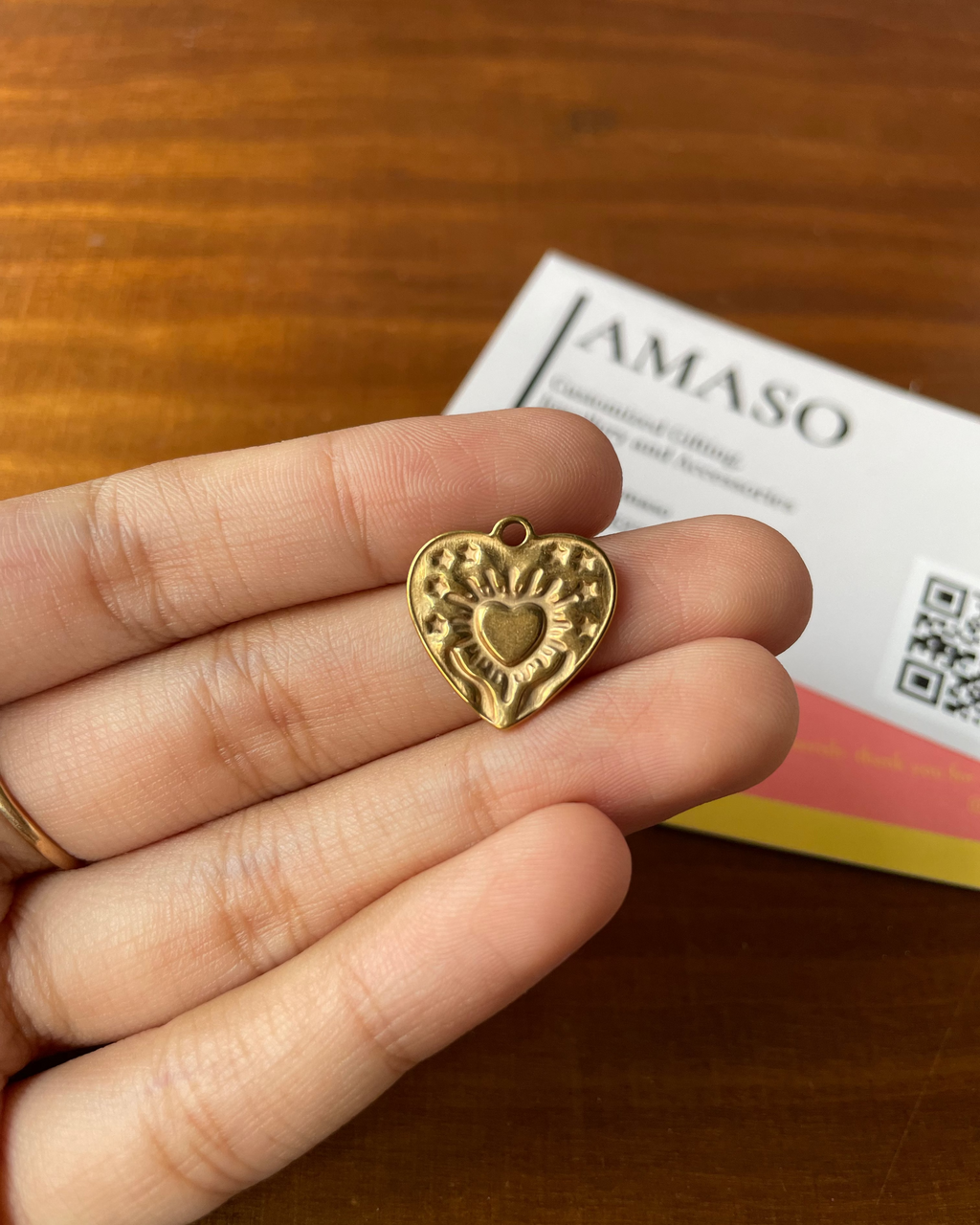 Gold heart-shaped pendant held between fingers with a 'JAMASO' card in the background.