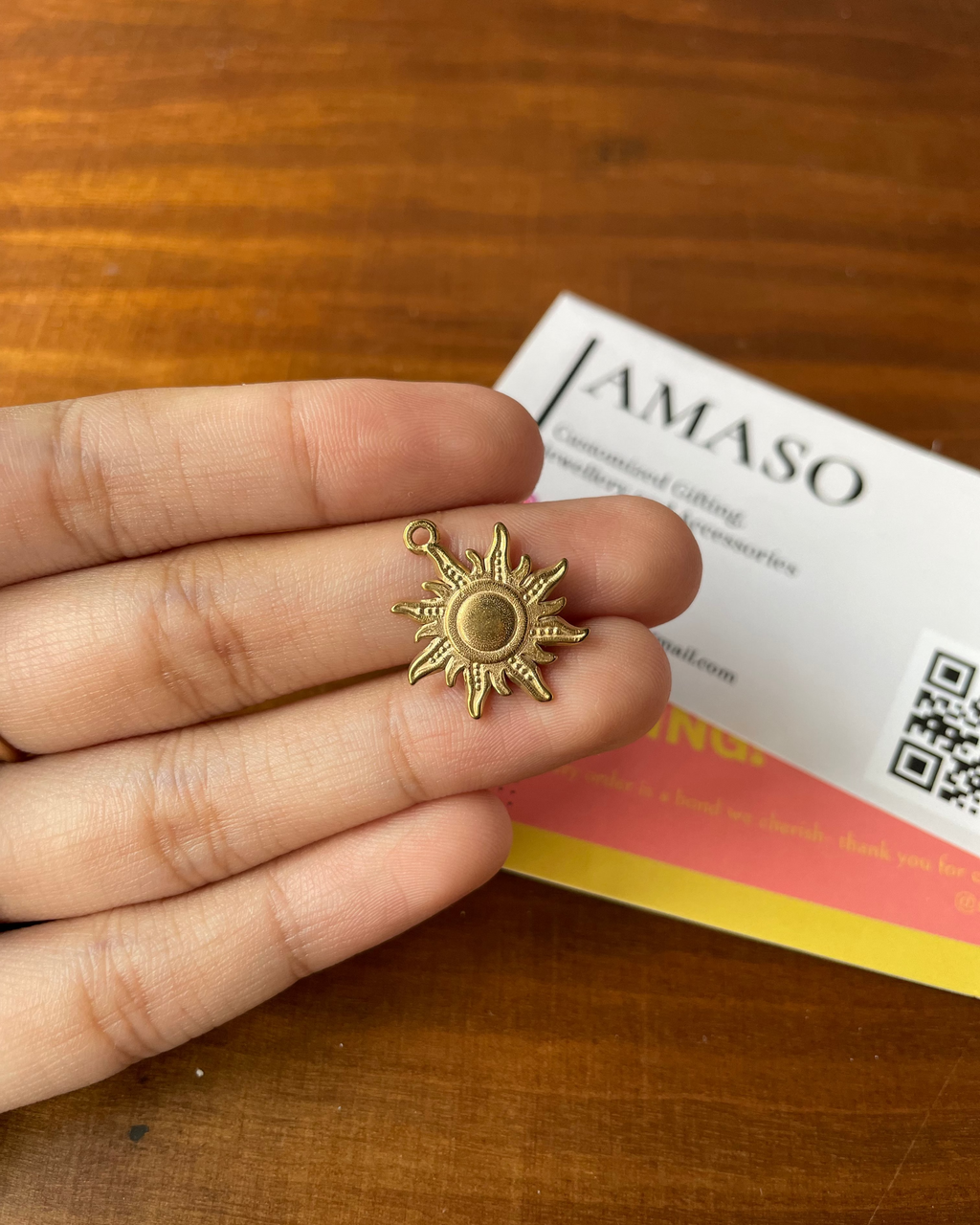 Gold sun-shaped earring held between fingers on a wooden surface with a JAMASO product in the background.