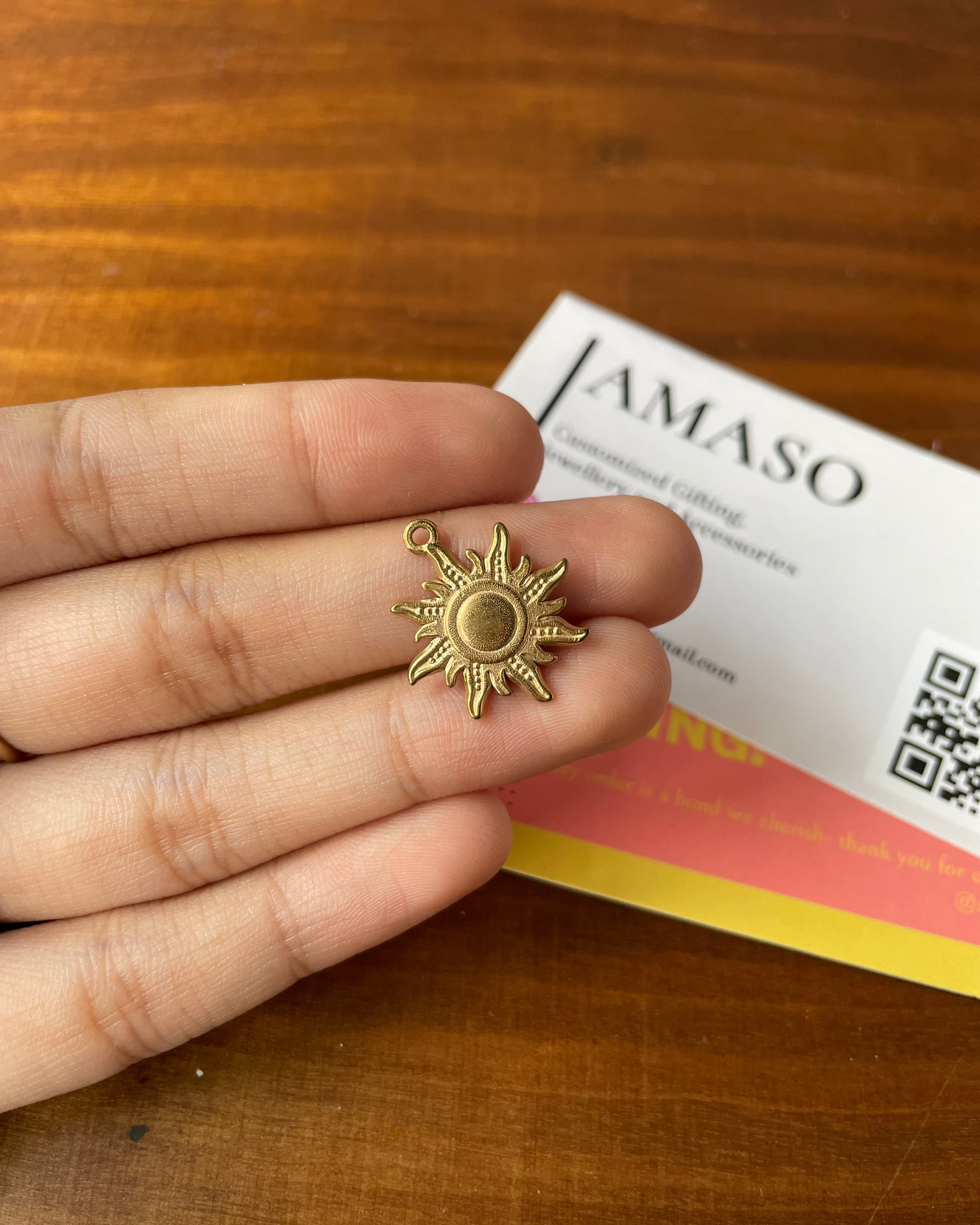 Gold sun-shaped earring held between fingers on a wooden surface with a JAMASO product in the background.