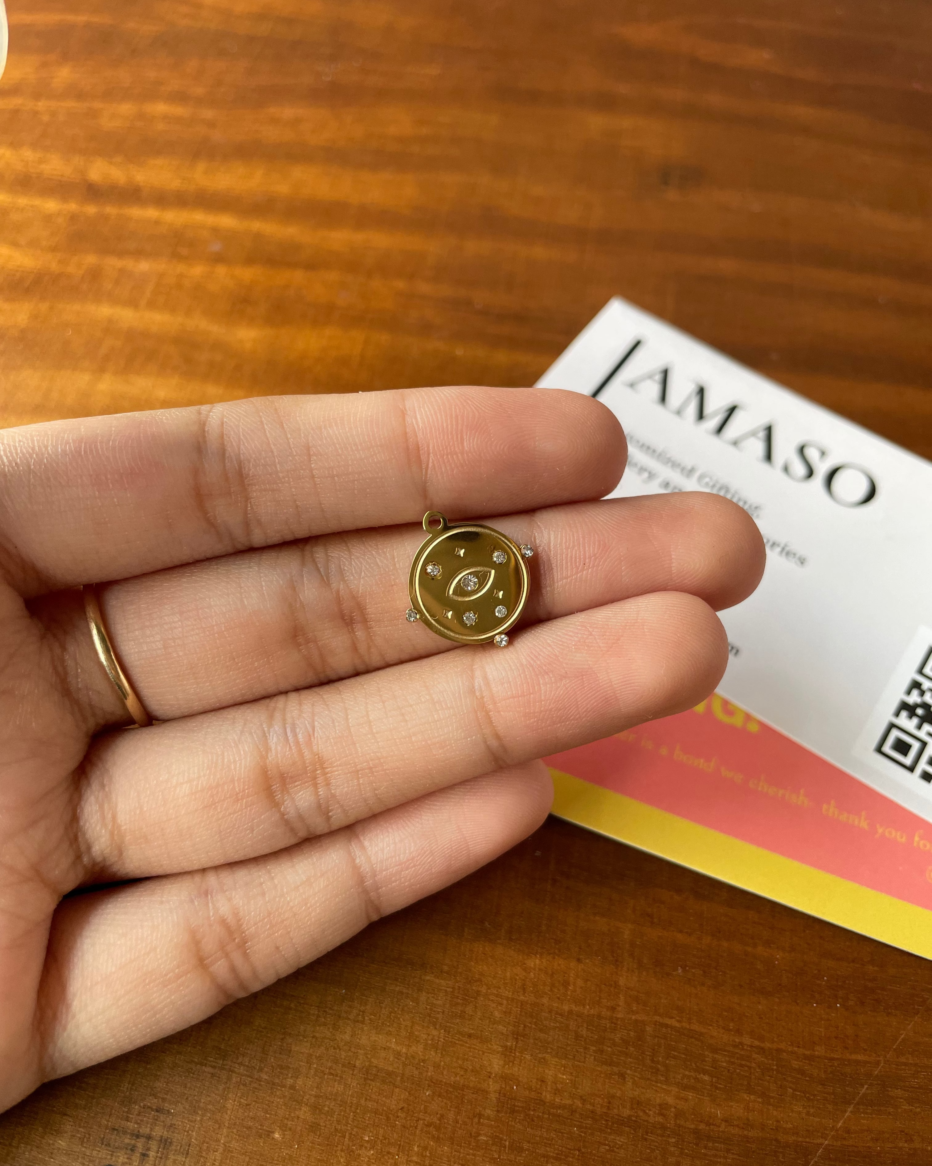 A close-up of a hand holding a small gold compass charm, with a wooden table background and a branded card partially visible behind it.
