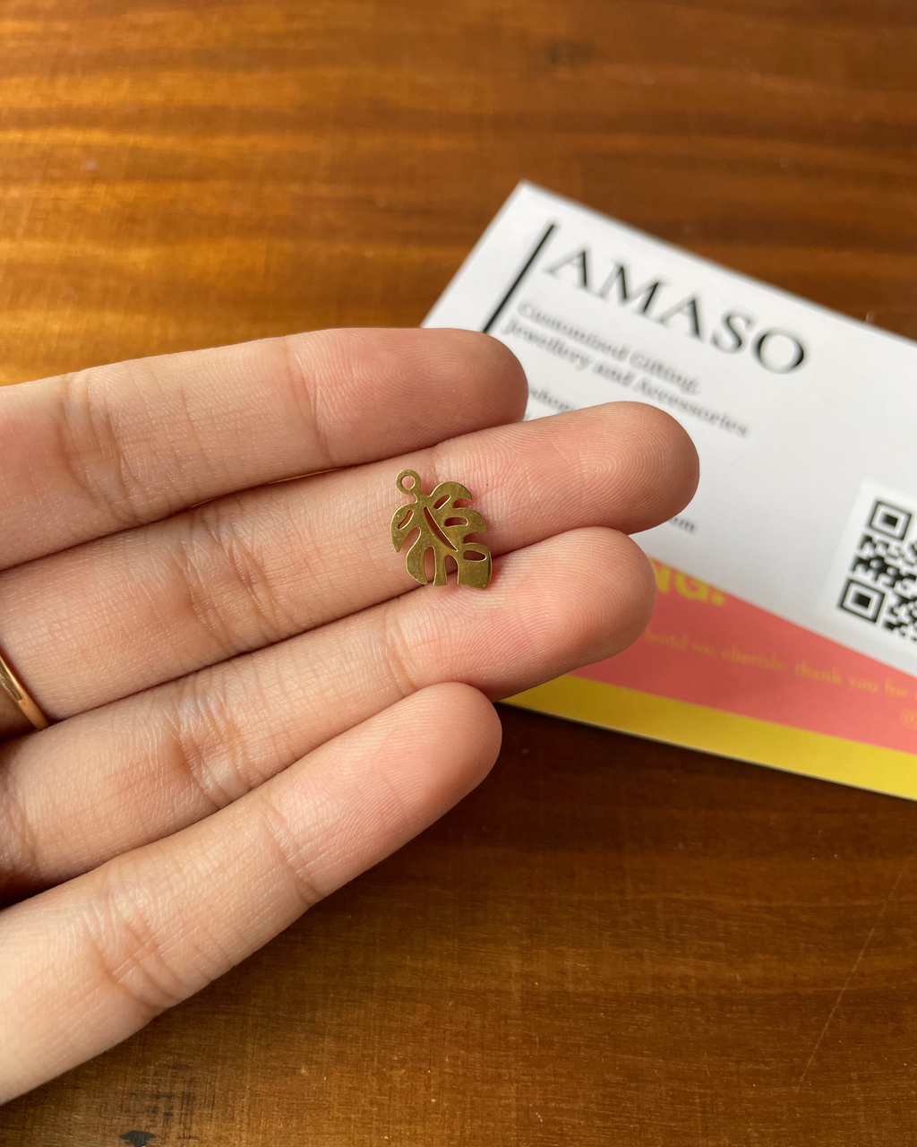 Gold leaf-shaped earring held between fingers on a wooden surface with AMASO branding in the background.
