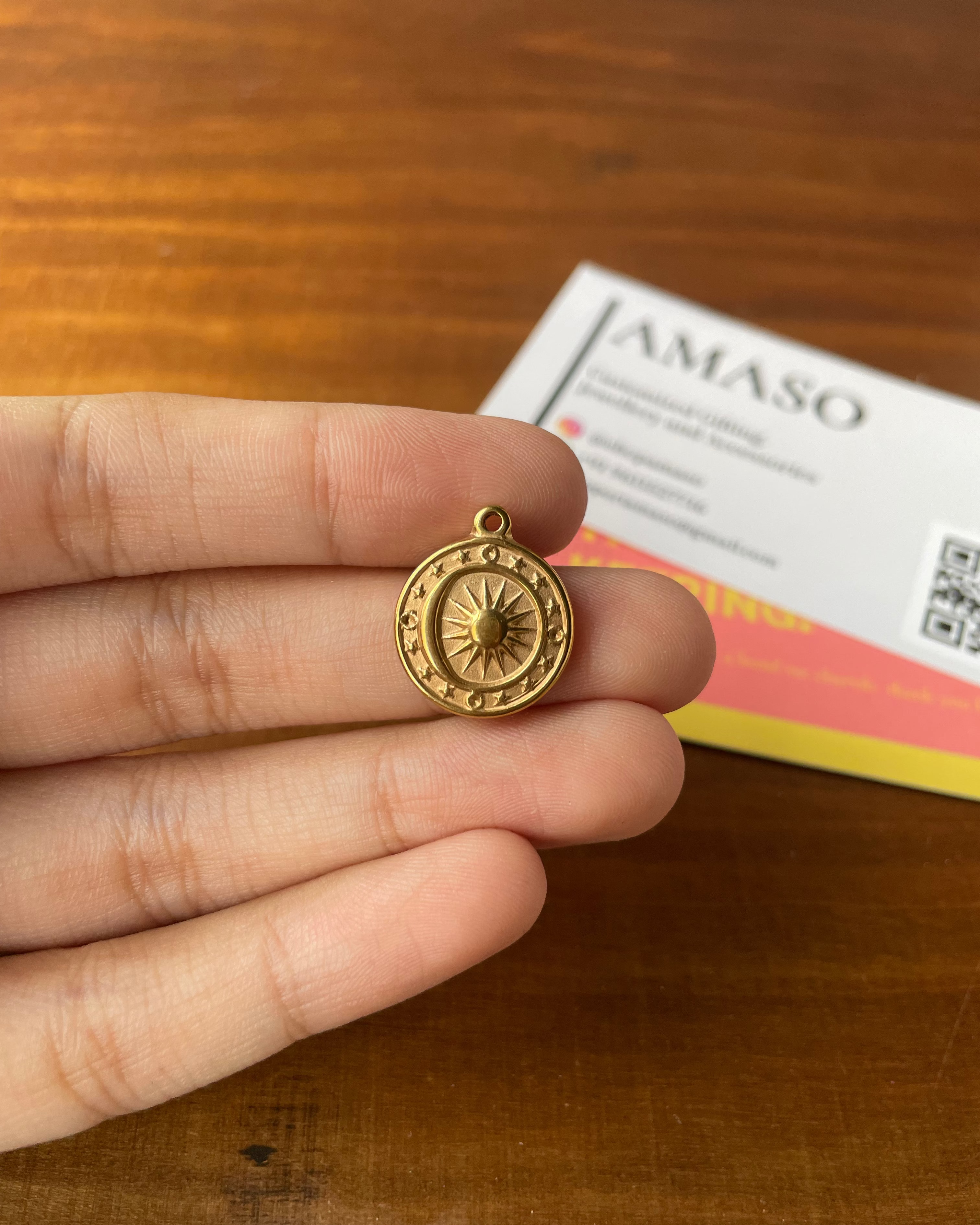 A small round gold pendant with a sunburst design in the center, held between fingers over a wooden table, with a blurred card in the background.