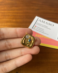 A hand holds a small gold pendant with a colorful floral design and a clear stone, with a branded card in the background on a wooden surface.
