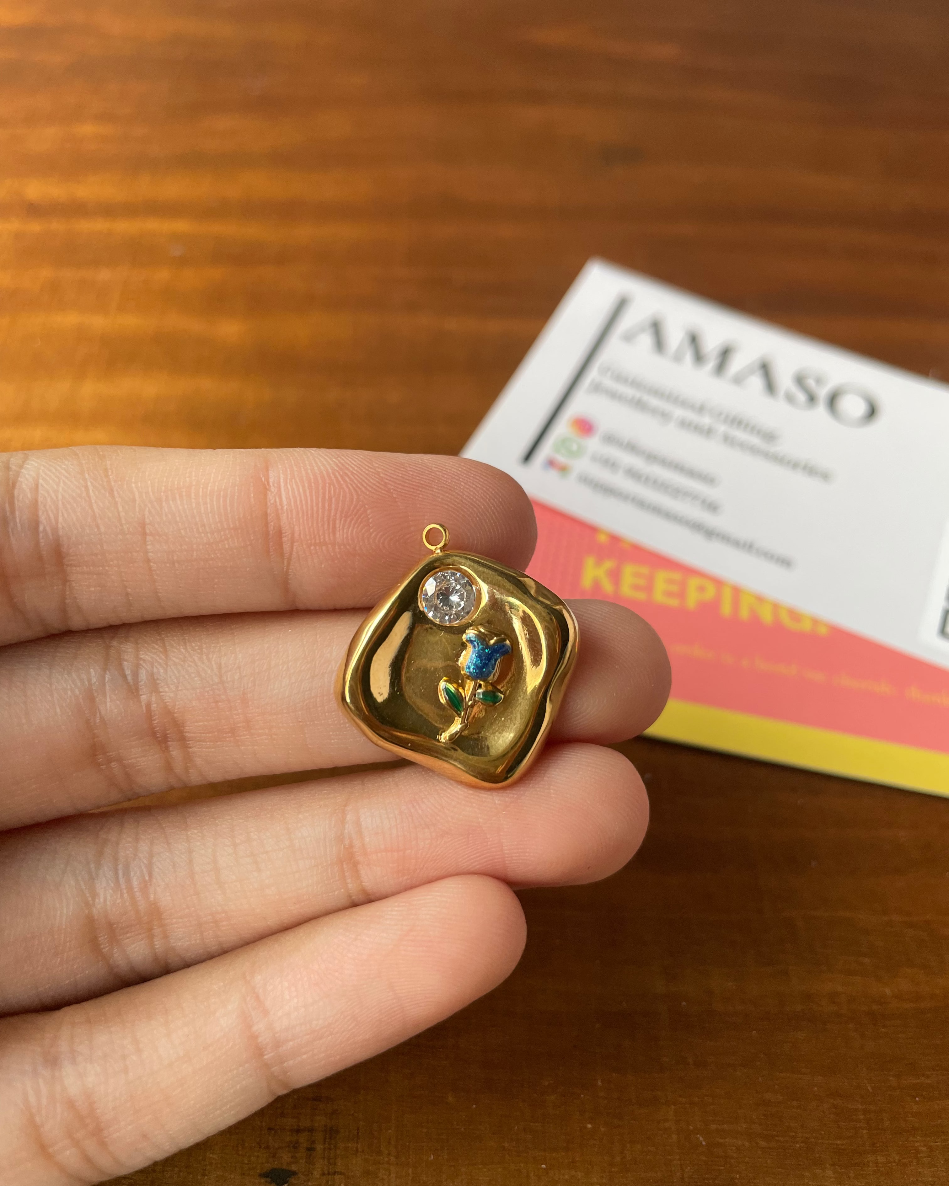 A hand holds a small gold pendant with a colorful floral design and a clear stone, with a branded card in the background on a wooden surface.