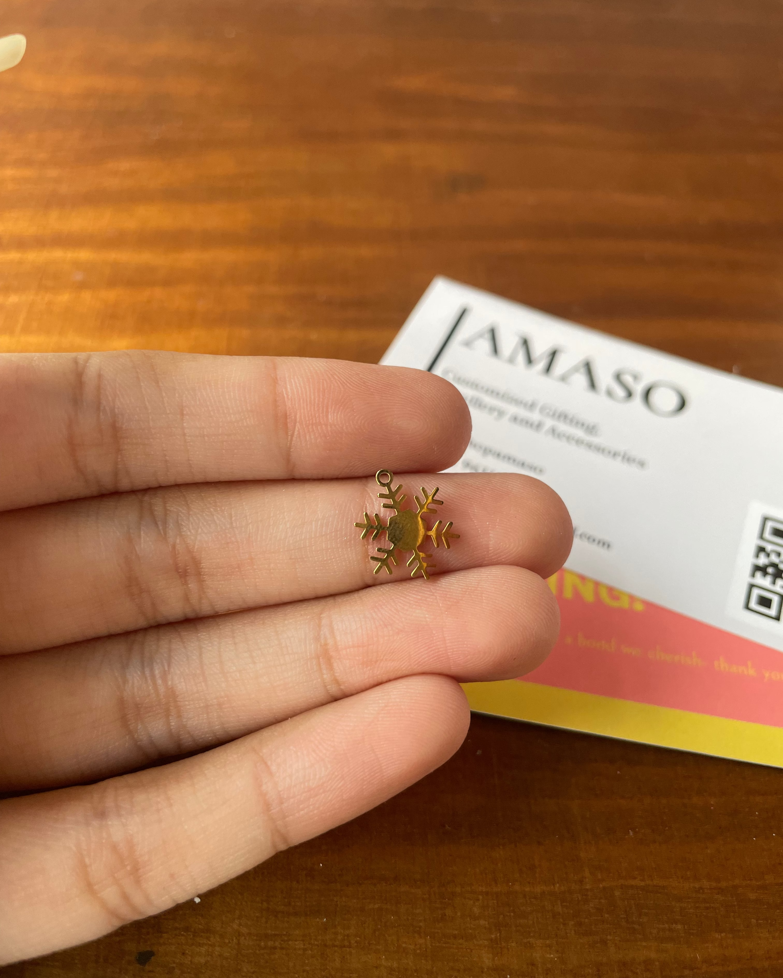 Small gold snowflake charm held between fingers with a 'AMASO' card in the background on a wooden surface.