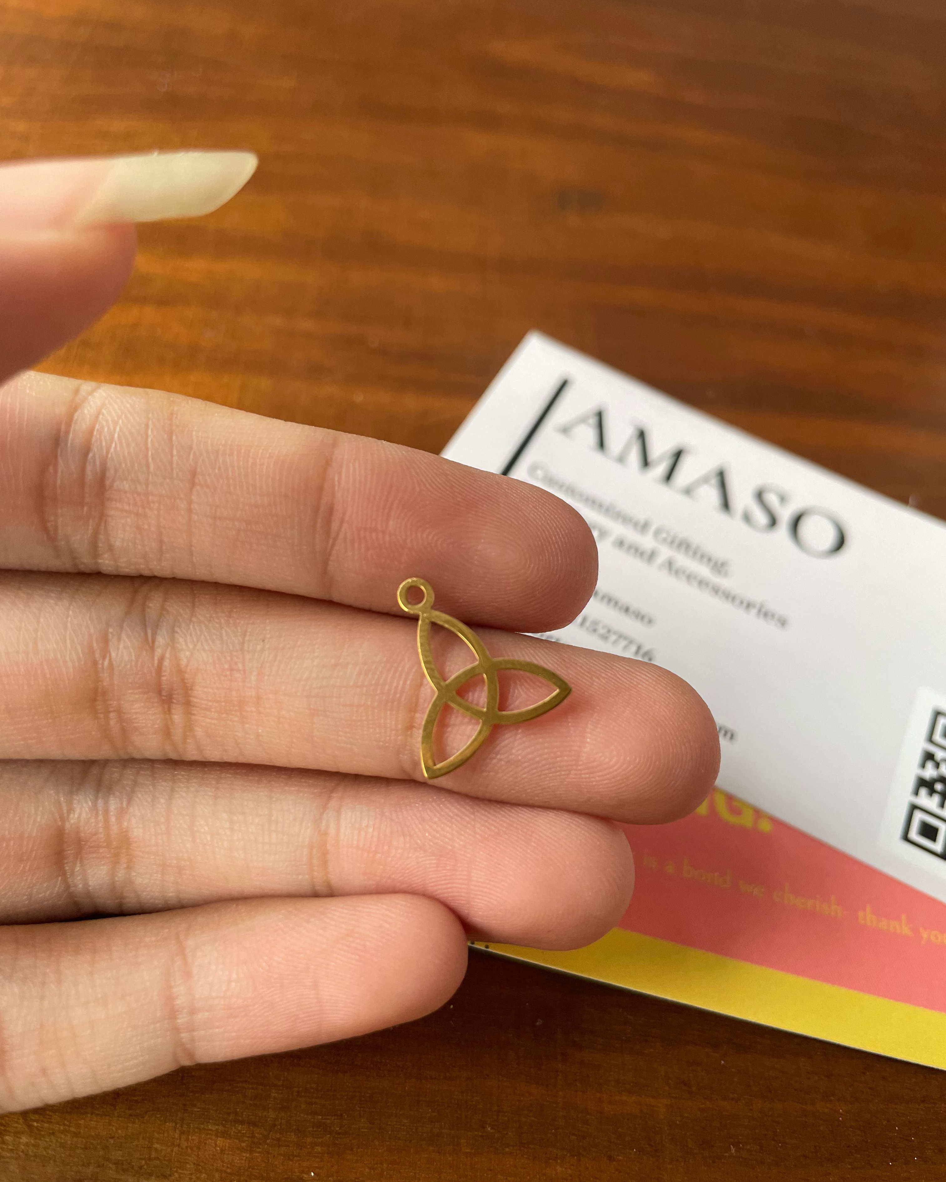 Gold Trinity knot pendant held between fingers with AMASO card in background
