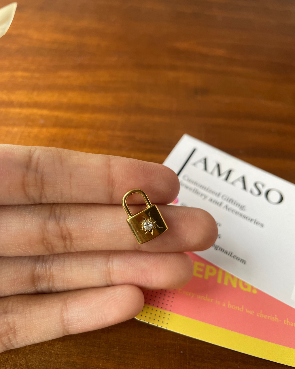 Gold lock-shaped earring held between fingers on a wooden surface with a JAMASO card in the background.