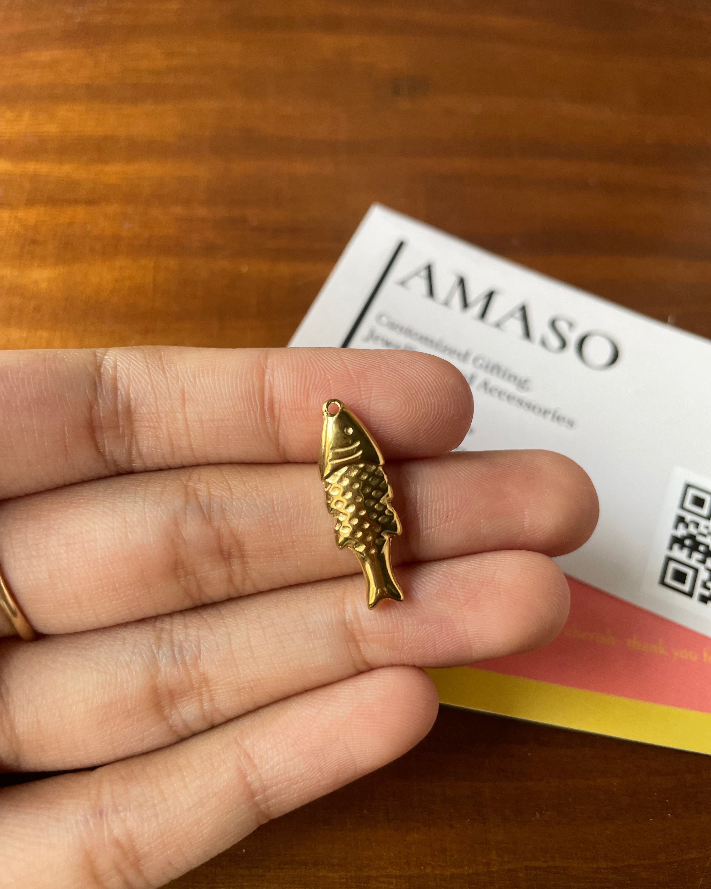 Gold fish-shaped pendant held between fingers with a 'AMASO' card in the background.