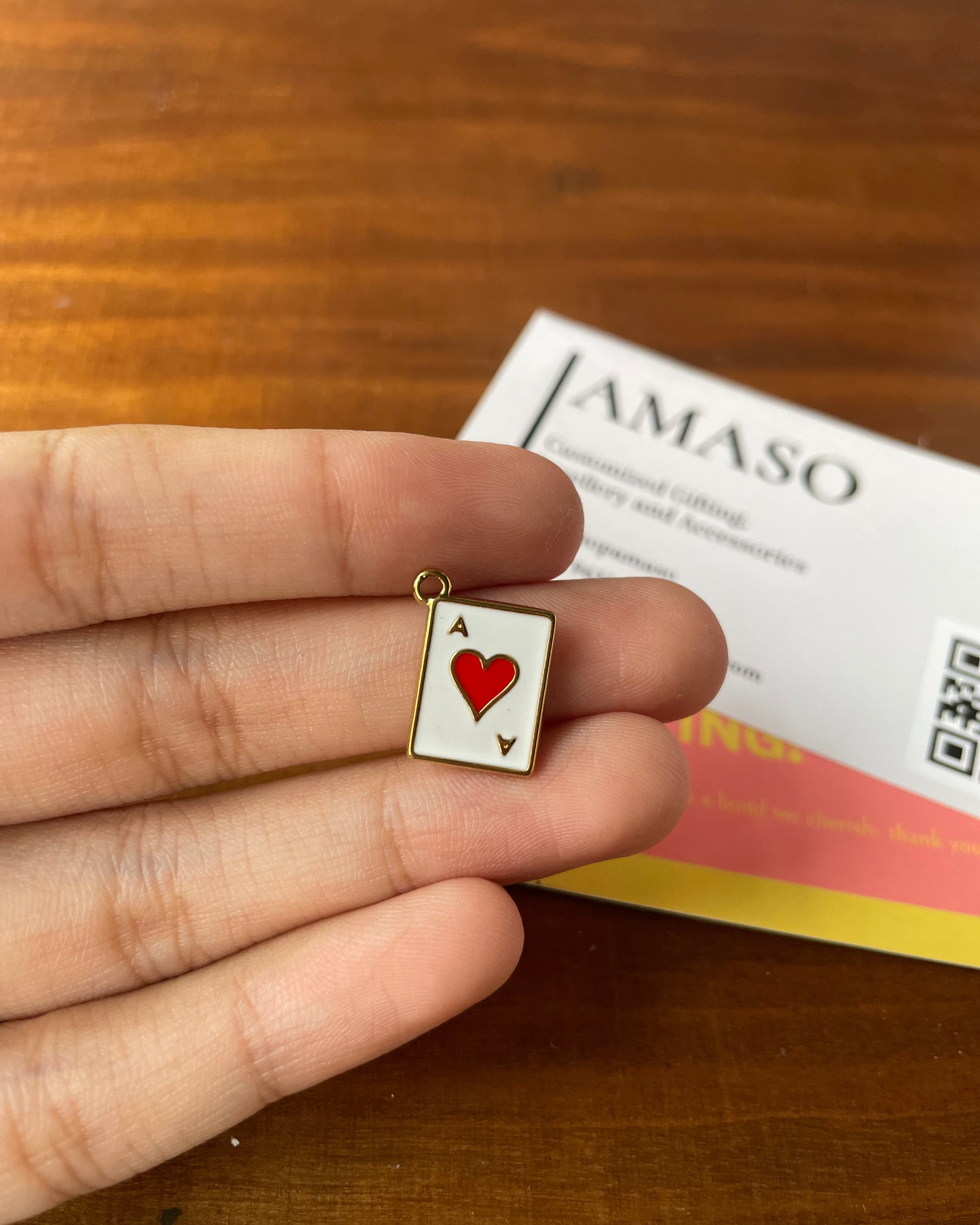 Hand holding a small heart-shaped card charm with a 'JAMASO' product in the background.