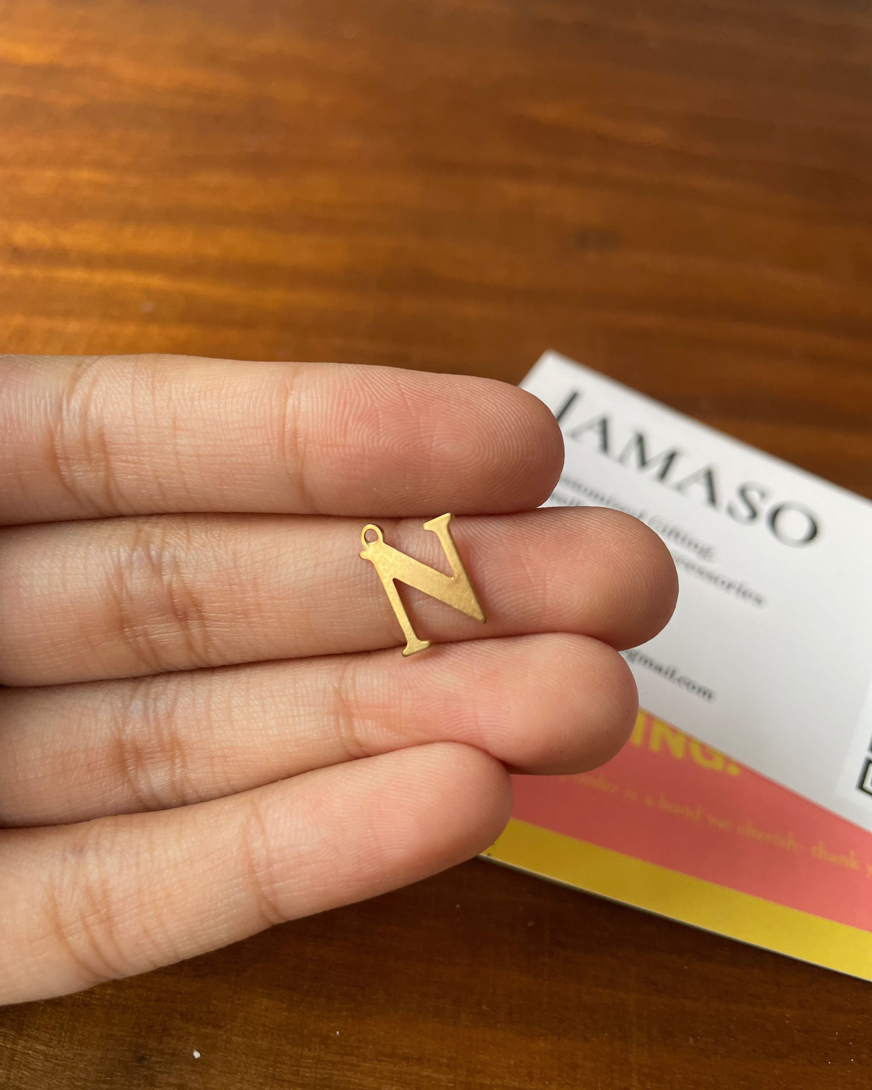Gold letter 'N' earring held between fingers on a wooden surface with a book in the background.