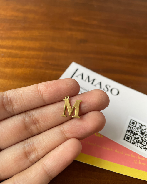 Gold letter 'M' charm held between fingers with Jamaso product in the background