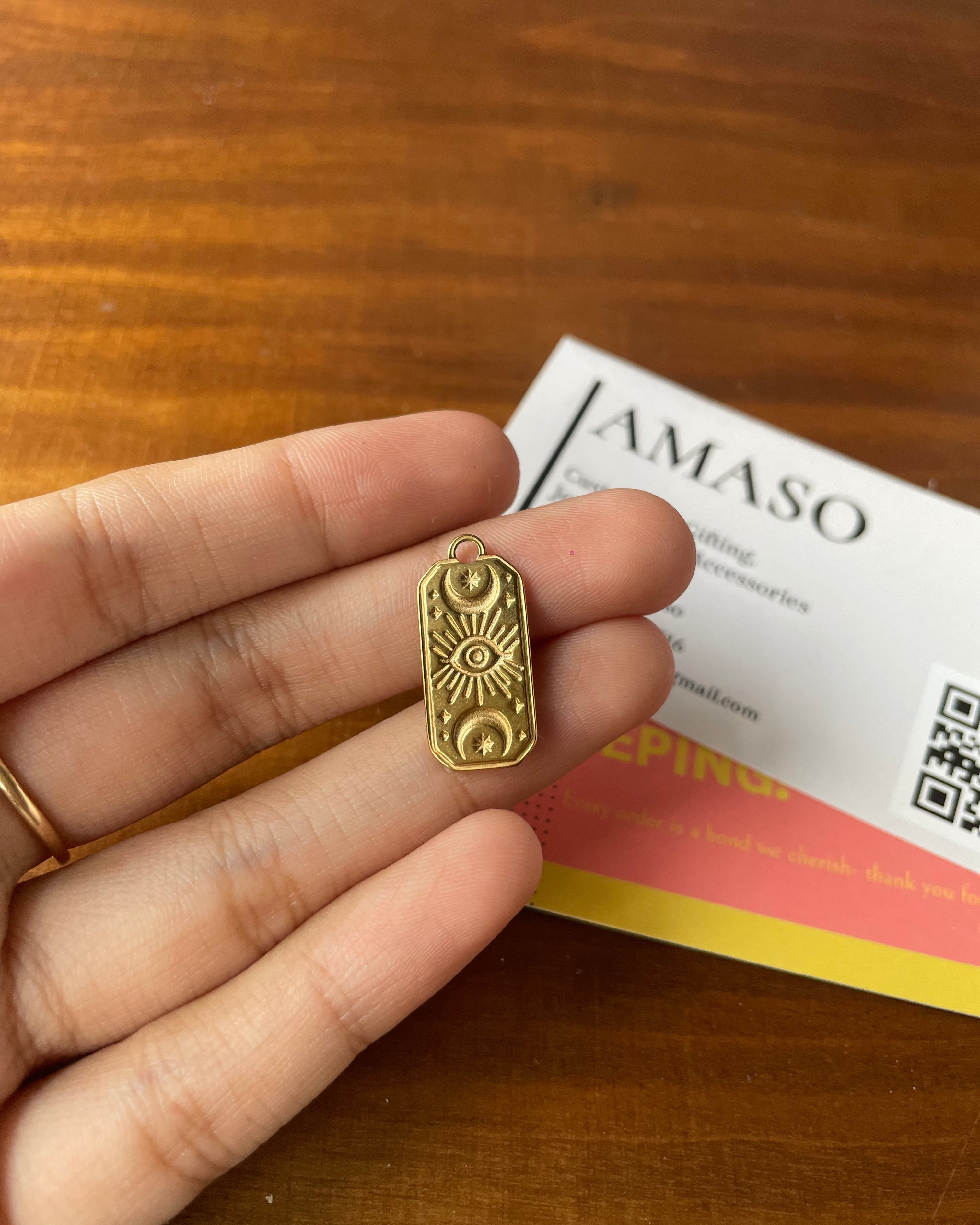 Gold pendant held between fingers with a wooden background and AMASO card.