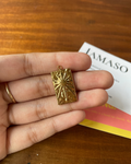 A rectangular gold pendant with a sunburst design is held on a person's fingers over a wooden table, with a branded card partially visible in the background.