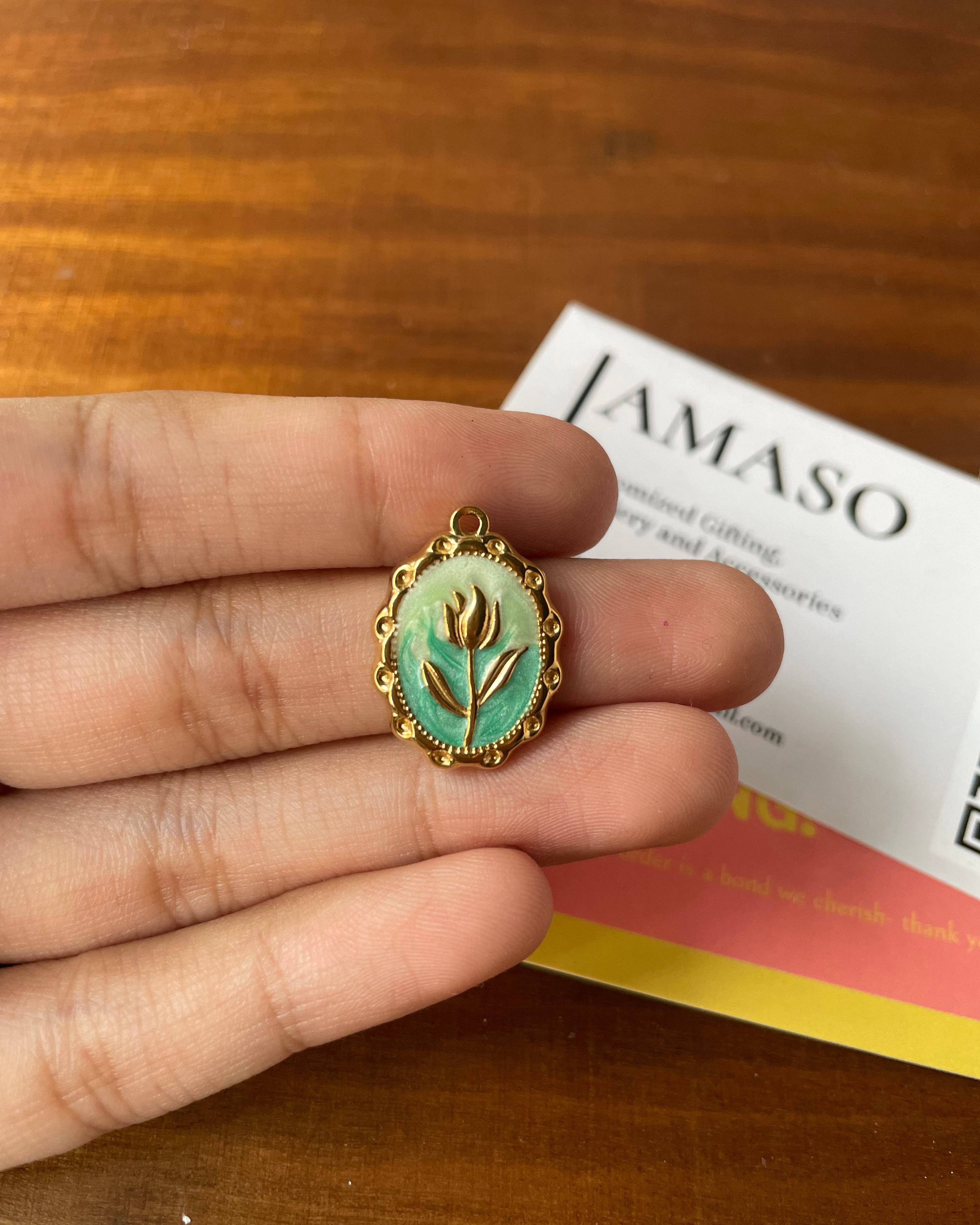 Gold pendant with a green stone and floral design held between fingers on a wooden surface with 'Jamaso' branding.