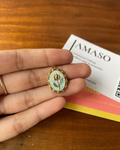 Hand holding a small decorative pendant with a leaf design on a wooden surface with AMASO branding.