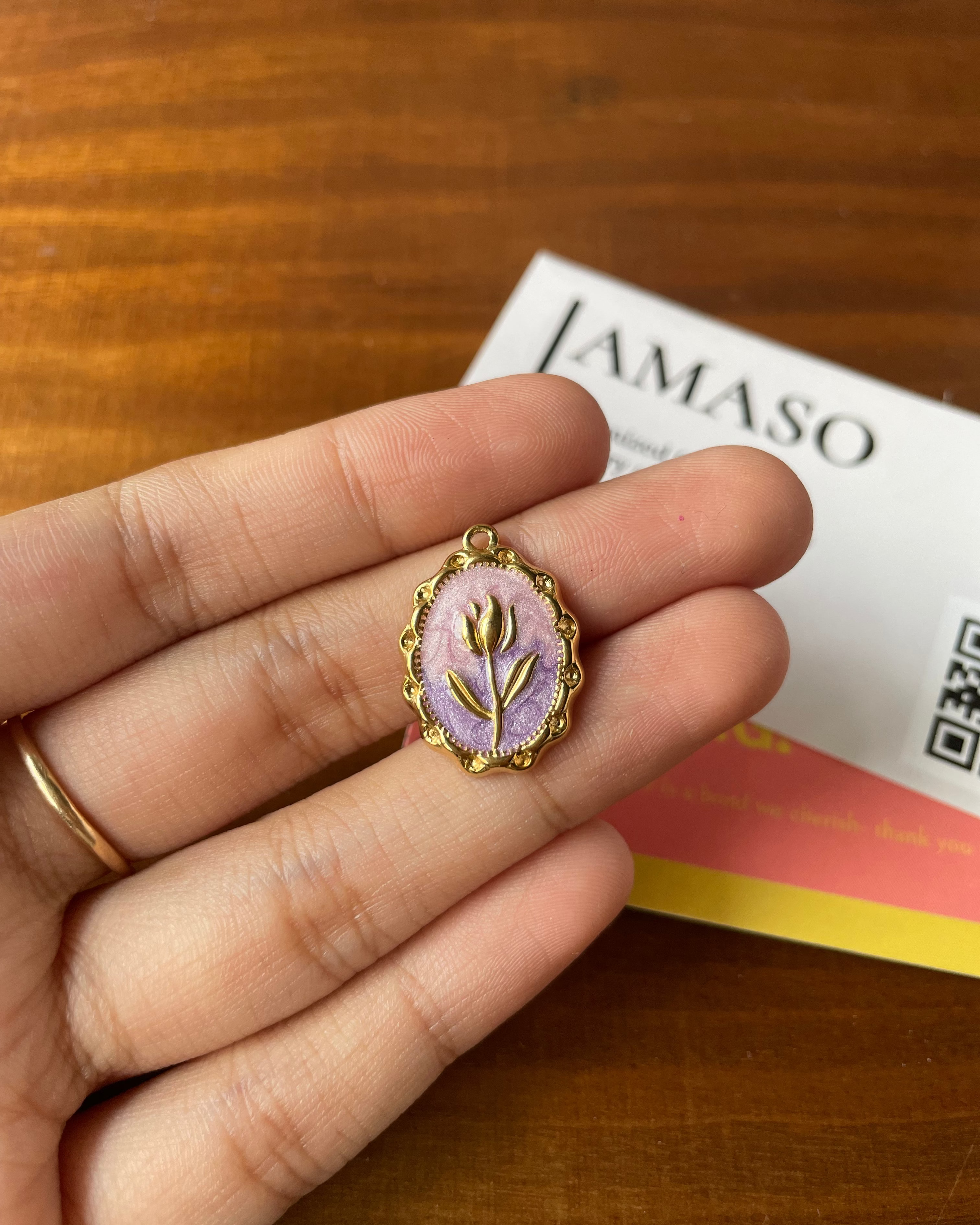 Hand holding a small gold pendant with a floral design on a wooden surface with a card in the background.