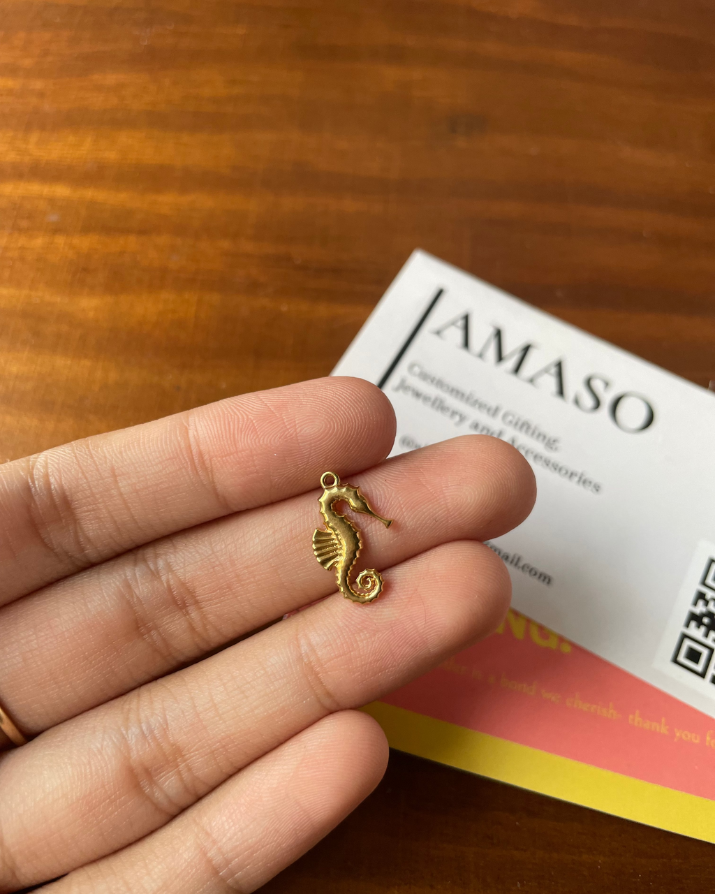 Gold seahorse charm held between fingers with AMASO brand card in the background