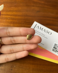 Hand holding a small gold shell-shaped earring with a JAMASO card in the background on a wooden surface.
