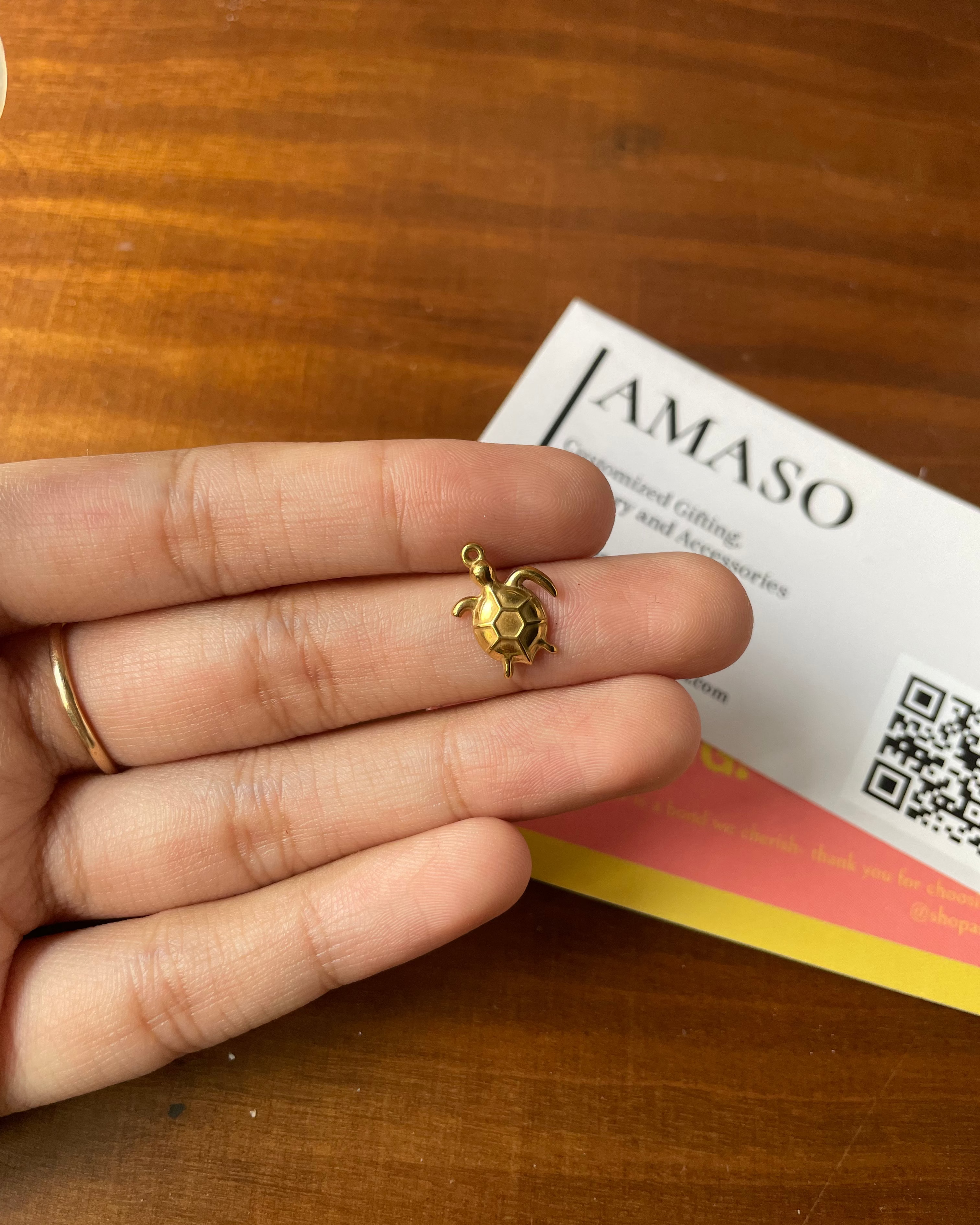 Gold turtle pendant held between fingers on a wooden surface with a 'JAMASO' card in the background.