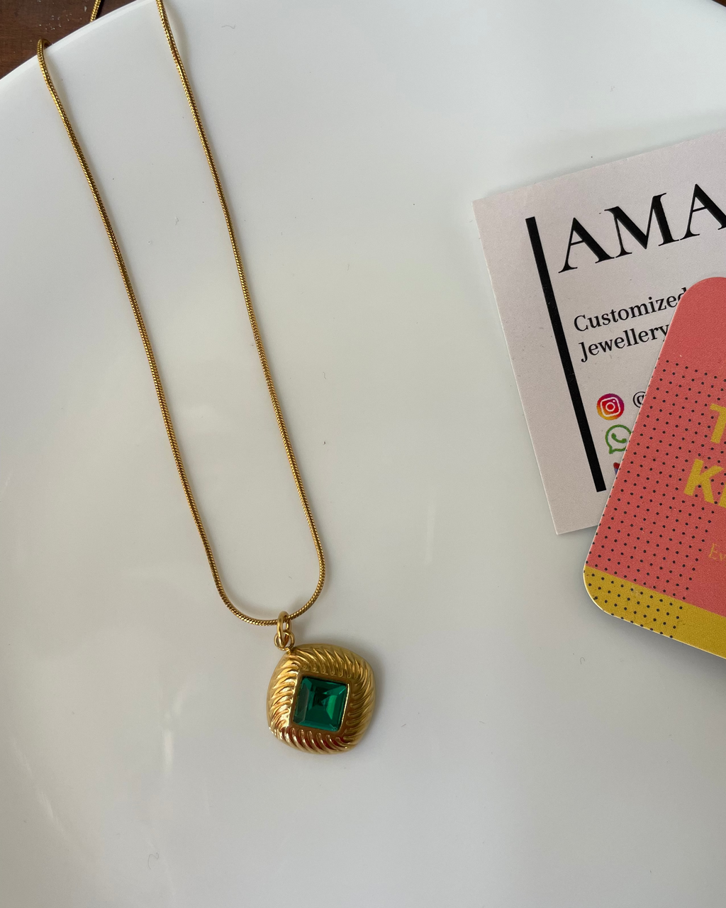 Gold necklace with a green pendant on a white surface next to a pink card with text.