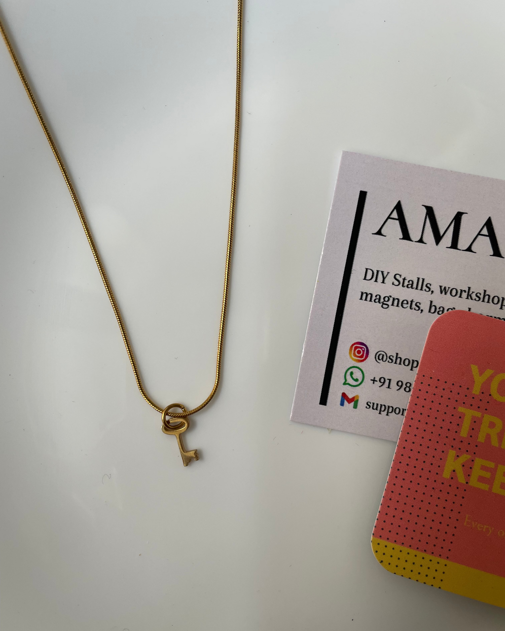 Gold necklace with a key pendant on a white surface next to a business card.