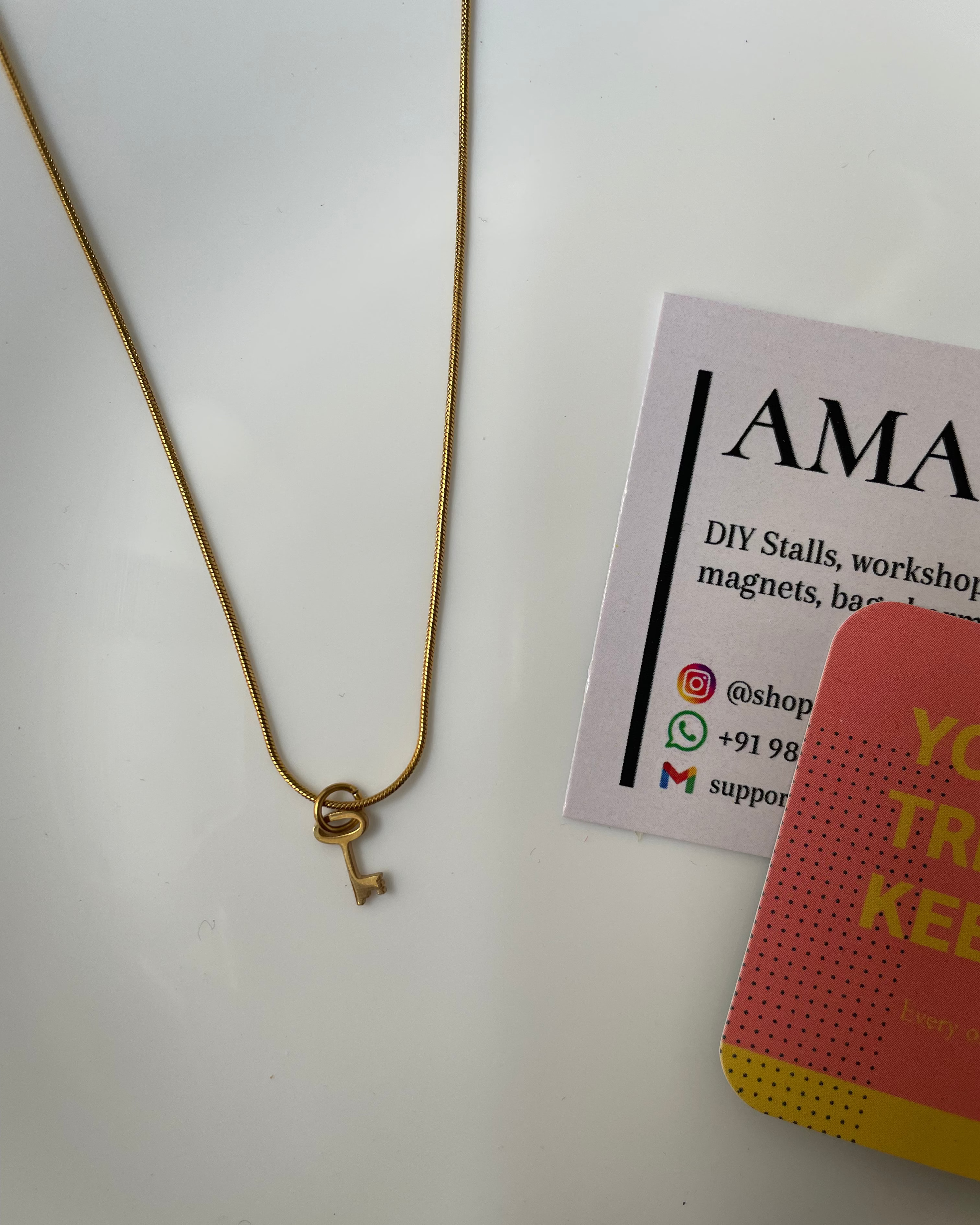 Gold necklace with a key pendant on a white surface next to a business card.