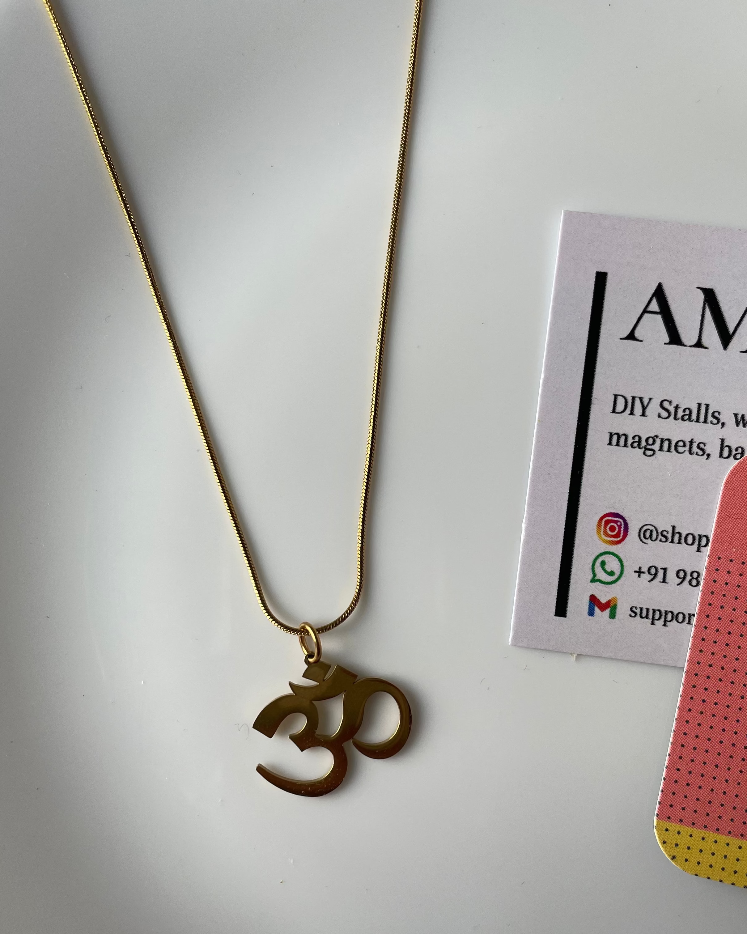 Gold necklace with Om pendant on a white surface with AMAS branding.