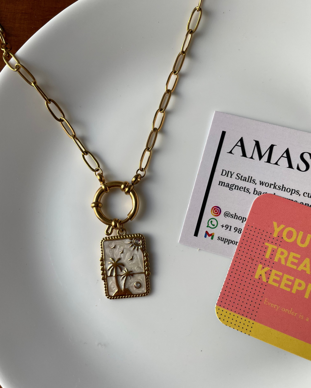 Gold necklace with a pendant on a white plate with a business card and pink card.