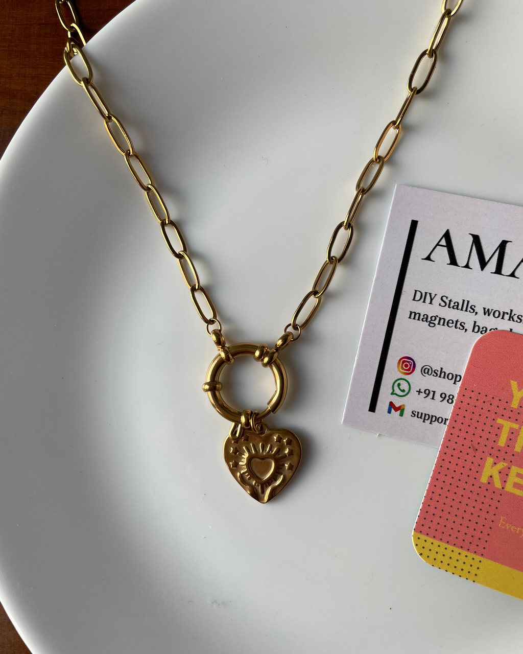 Gold necklace with a heart pendant on a white plate, next to an AMA branded card.