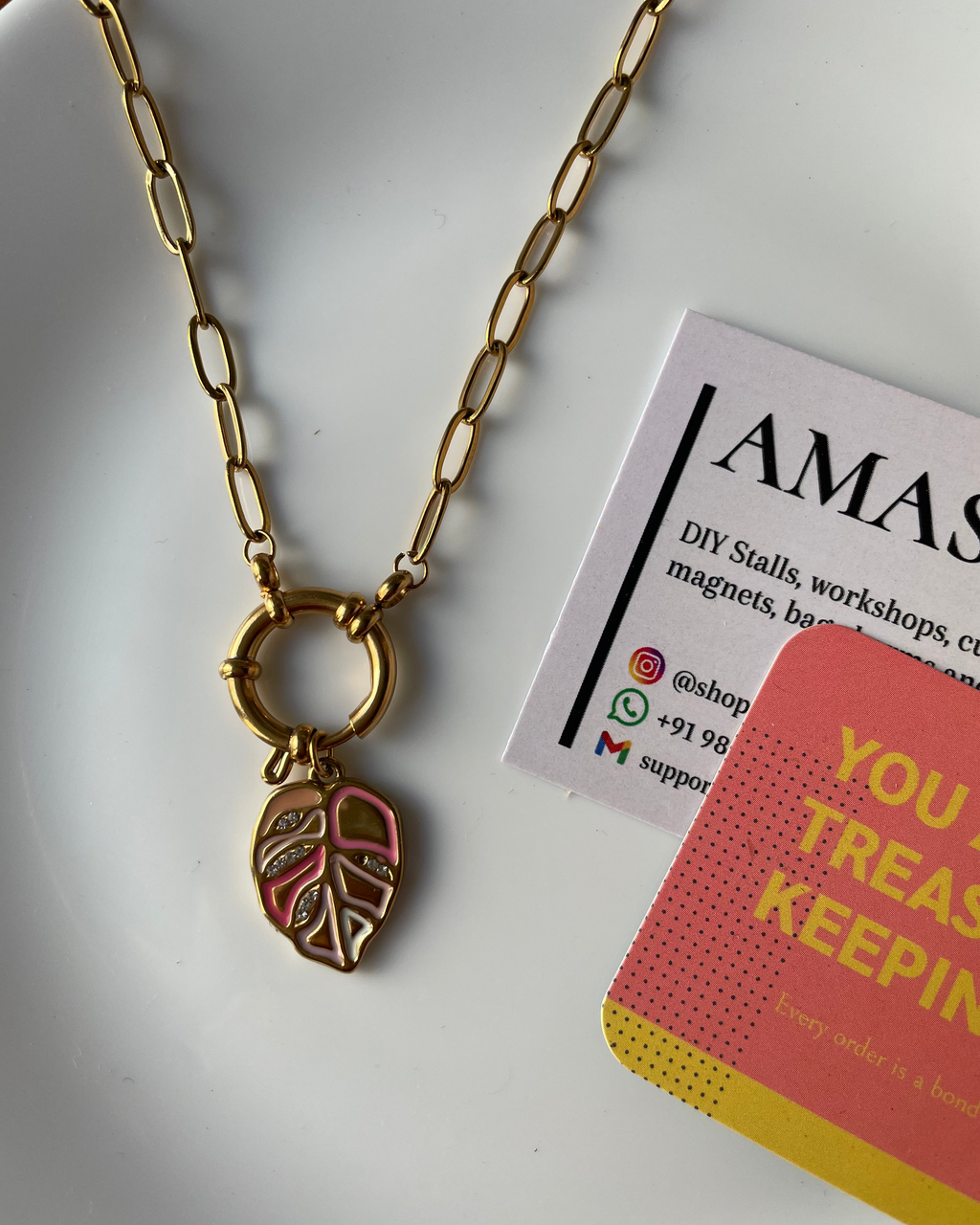 Gold necklace with a leaf charm on a white surface next to a business card and a colorful book.