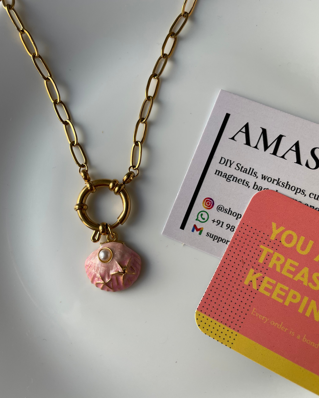 Gold chain necklace with a pink pendant on a white surface next to a business card and book.