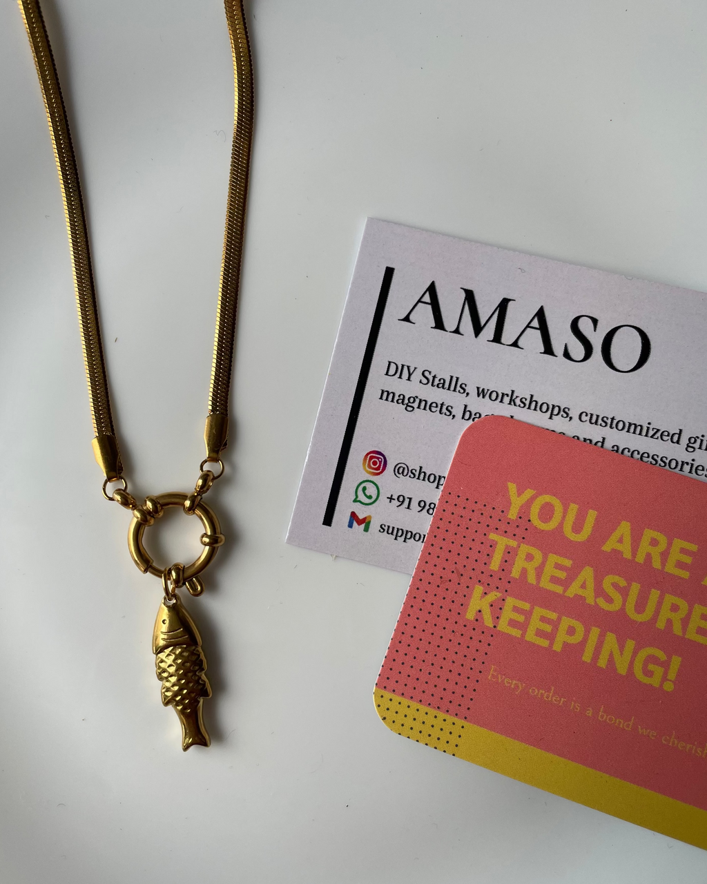 Gold necklace with fish pendant on a white surface next to AMASO branded materials.