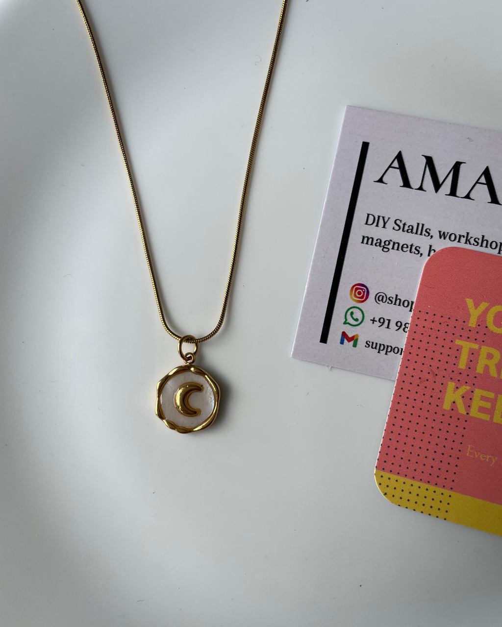 Gold necklace with a pendant on a white surface next to a business card.