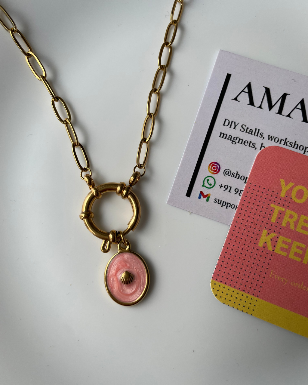 Gold chain necklace with a pink pendant on a white background, next to promotional materials.